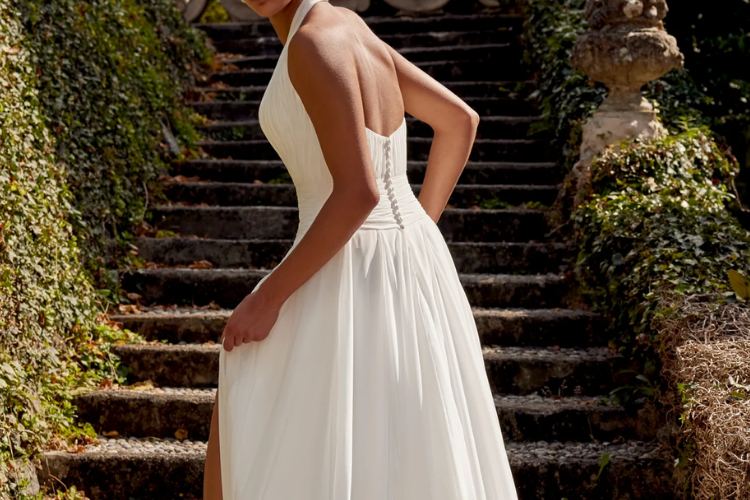 Bride in a halter-neck A-line wedding gown with a flowing skirt and thigh-high slit, posing on a stone staircase outdoors.
