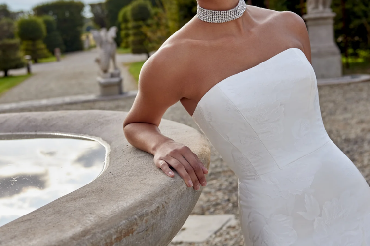 Close-up of a bride in a strapless wedding gown with subtle floral-textured fabric, leaning against a stone fountain outdoors.