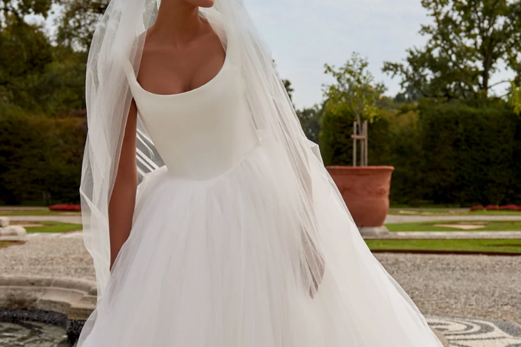 Bride in a scoop-neck ball gown with a tulle skirt and veil, standing outdoors in a garden setting.