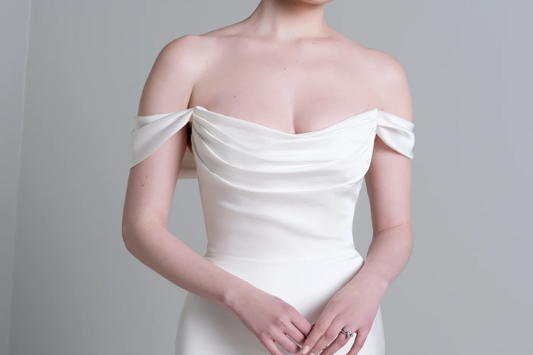 Bride wearing an off-the-shoulder wedding gown with a softly draped neckline and fitted silhouette against a neutral background.